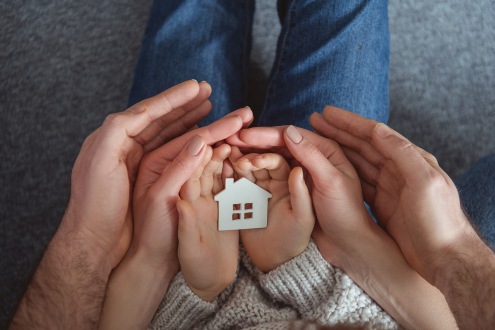A child has a paper house in their open hands. Two parents have their open hands surrounding the child's.