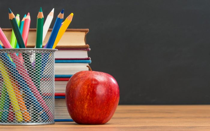 A cup of colored pencils, a stack of books and an apple on a desk.